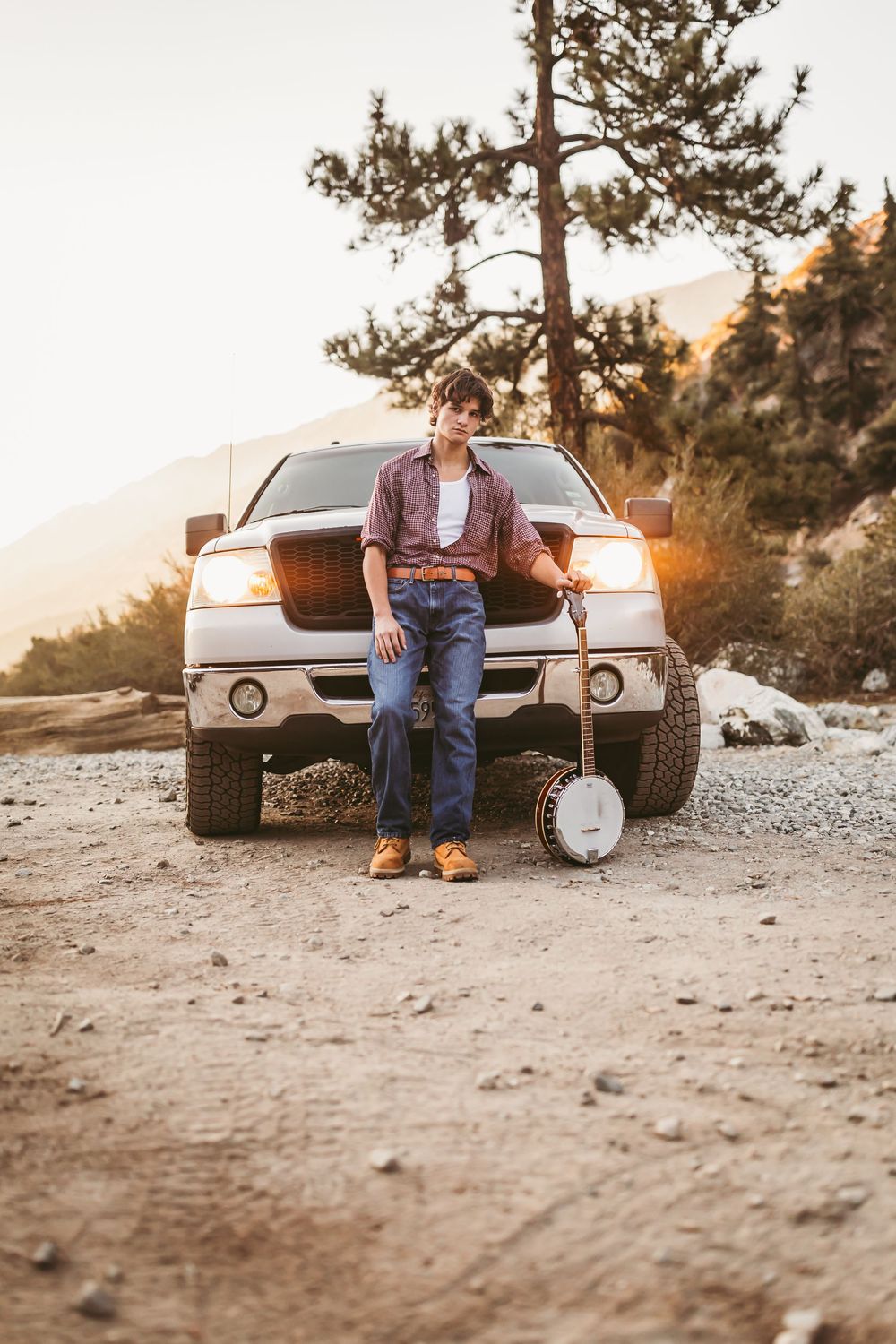 Young man leaning against white pickup truck holding banjo at golden hour in rural mountain setting.
