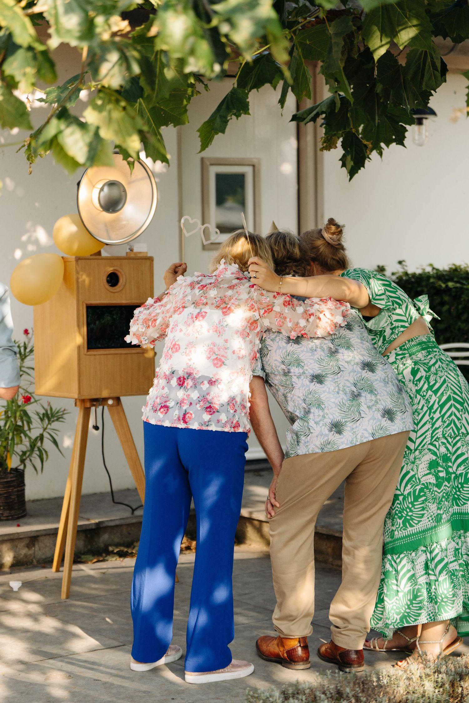 Three people posing together outdoors near vintage wooden photo booth at sunny garden party.