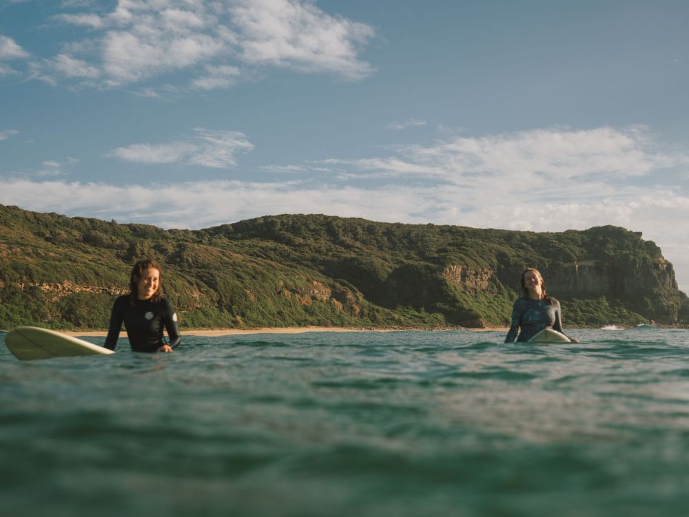two girls smiling in out of focus shot with cliffs behind with Sunlight surfers Newcastle women's surfing group at Dudley