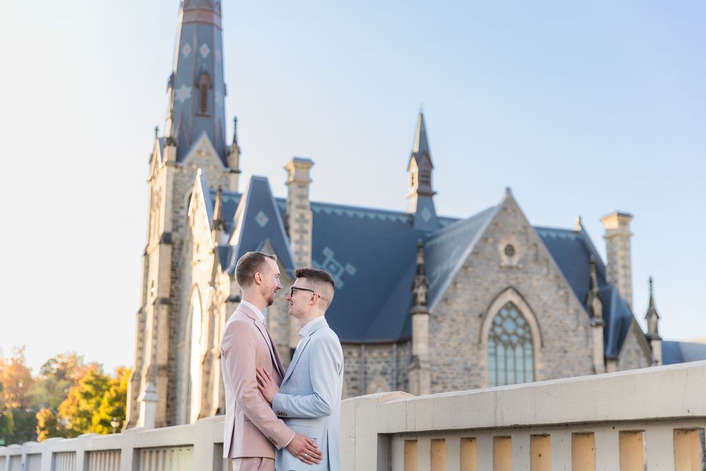 Groom and groom in pink and blue suits standing together on a bridge in front of a church downtown galt in Cambridge, Ontario.