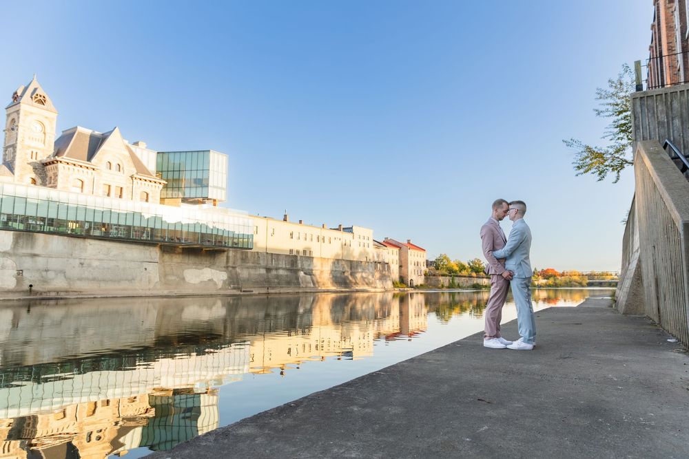 Two grooms in pink and blue suits stand close to each other beside the river during sunset downtown galt in Cambridge, Ontario.