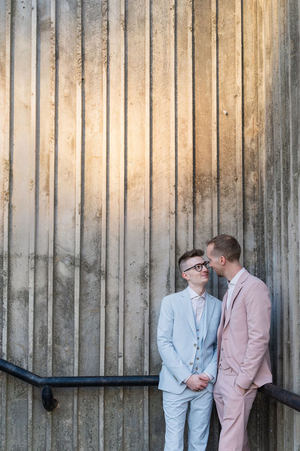 Groom and groom looking lovingly at each other in a stairwell wearing blue and pink suits by the river in downtown Galt, Cambridge, Ontario.