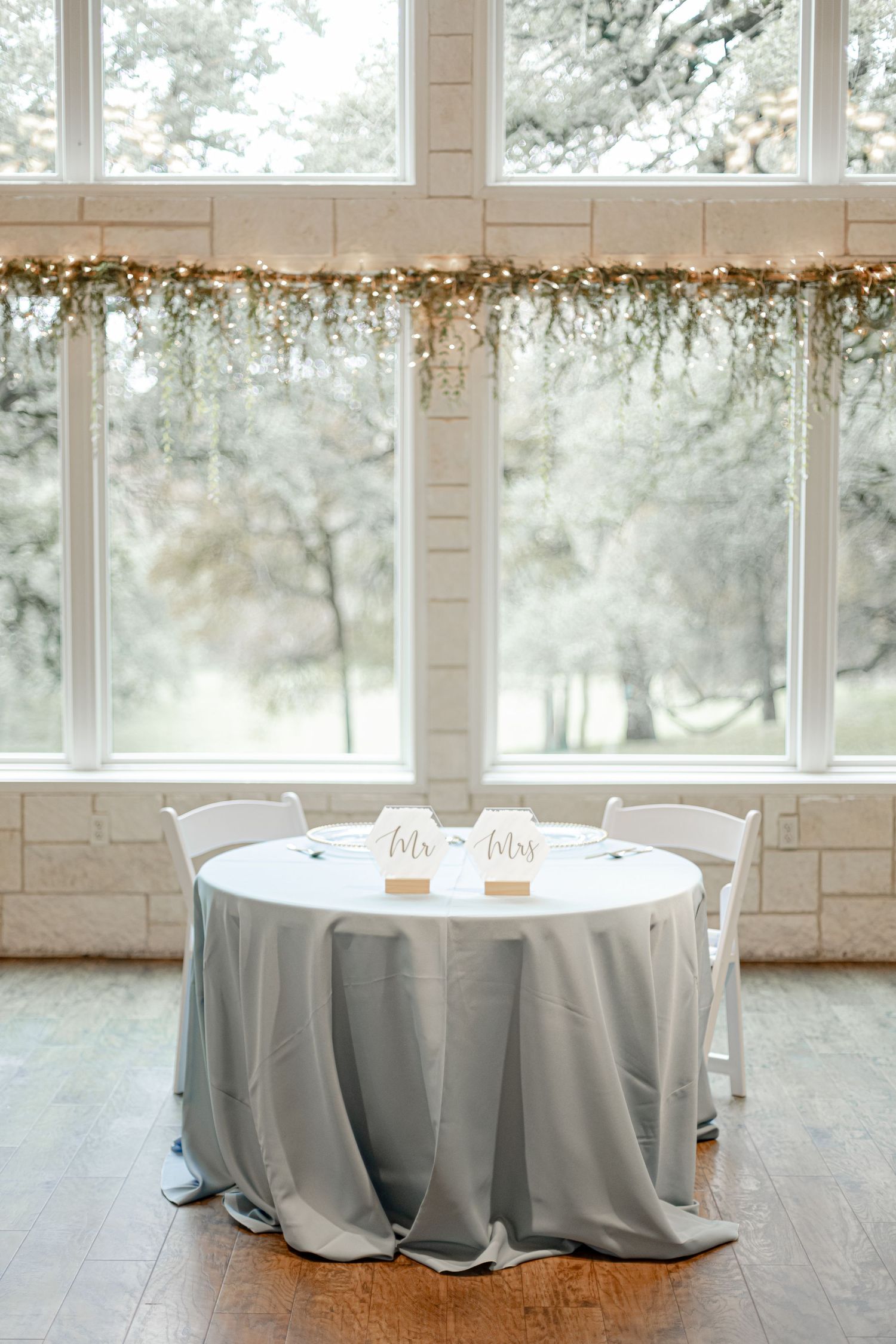 Elegant wedding sweetheart table with gray linen and Mr. Mrs. signs by large windows.