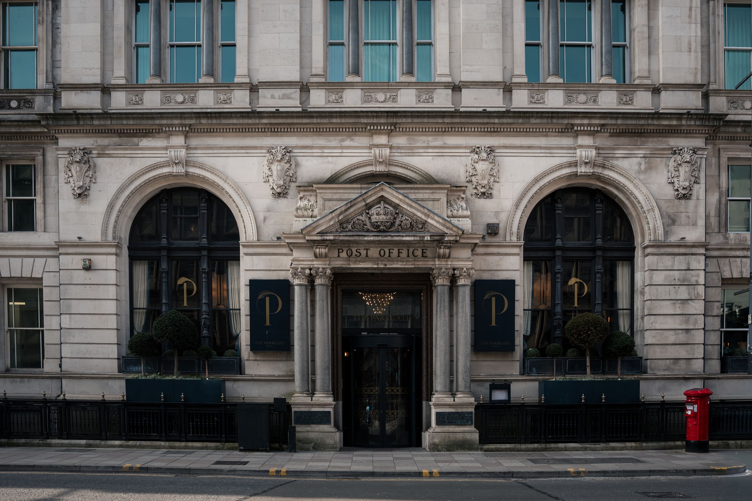 Grand neoclassical post office building facade with arched windows, ornate stonework, and a red postbox on a quiet street.