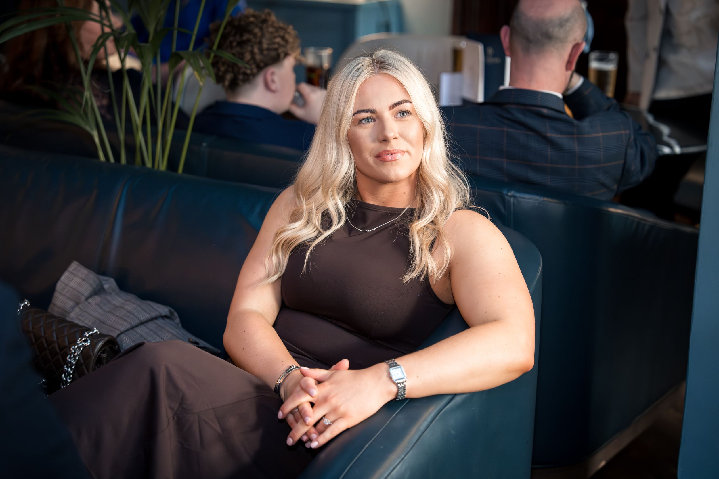 Blonde woman in brown sleeveless dress sits relaxed on blue leather sofa at an elegant venue.