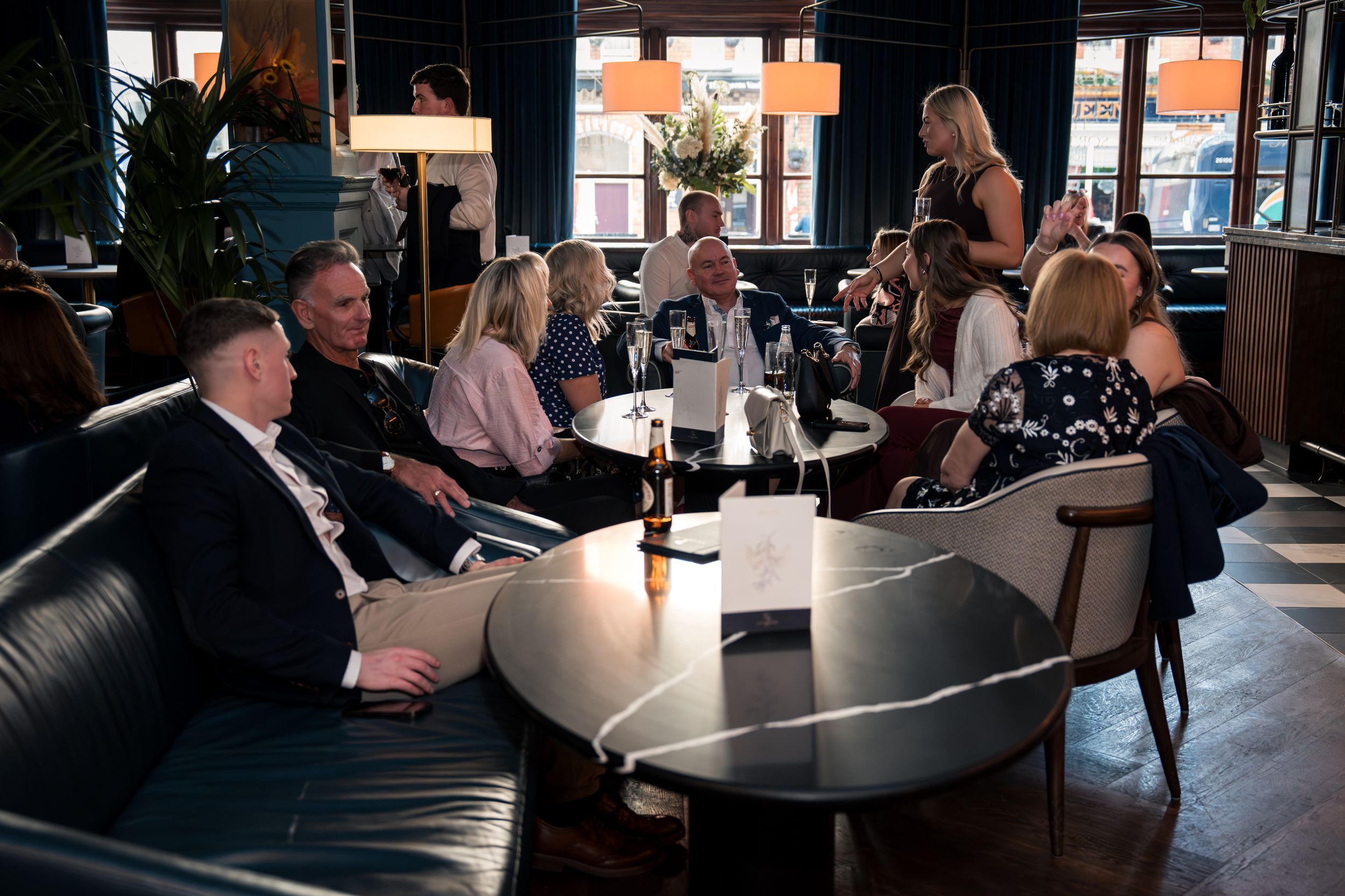 Group of adults socializing in a stylish dark blue bar lounge, seated on leather sofas around round tables.