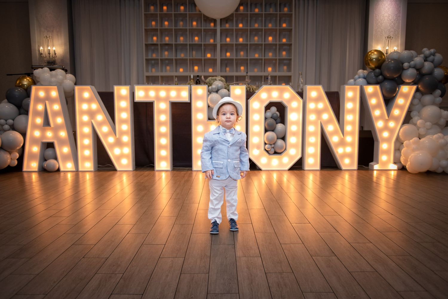A young boy in a blue suit stands before large illuminated marquee letters spelling ANTHONY at a party.