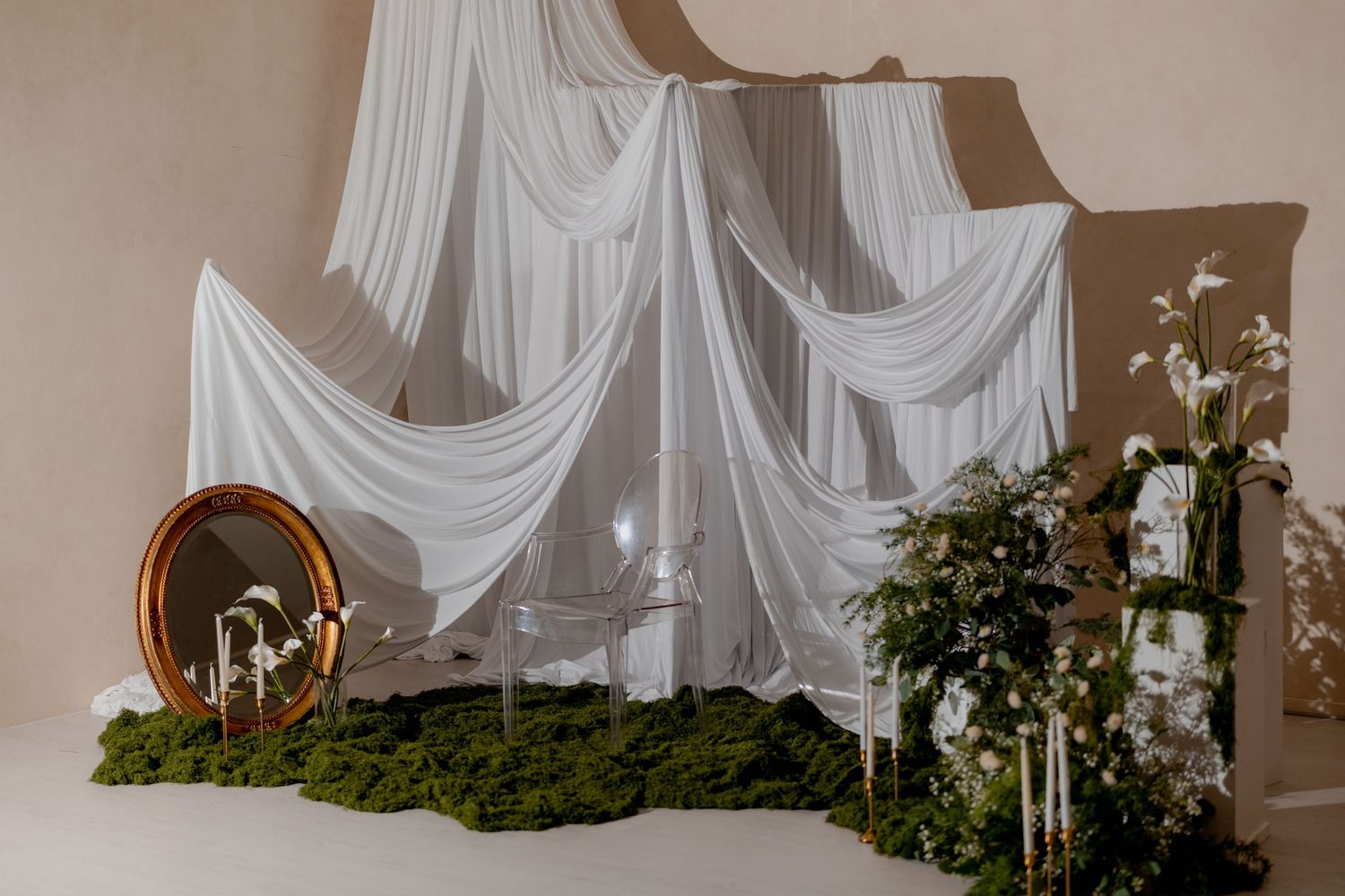 Elegant wedding backdrop with white draped fabric, moss ground cover, gold mirror, glass chair, and floral arrangements.