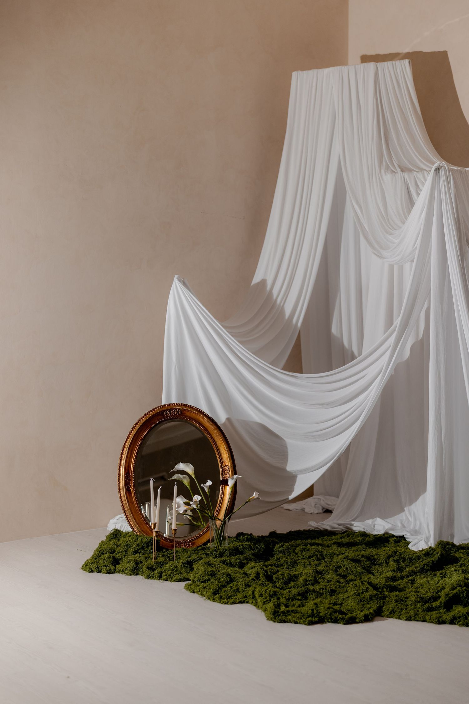 Elegant white draped fabric backdrop with a gold oval mirror, green moss, and candles on a beige studio floor.