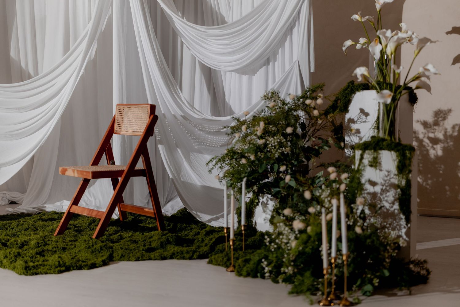 Elegant wedding ceremony backdrop with white draped fabric, moss floor, white candles, and floral arrangements featuring calla lilies.