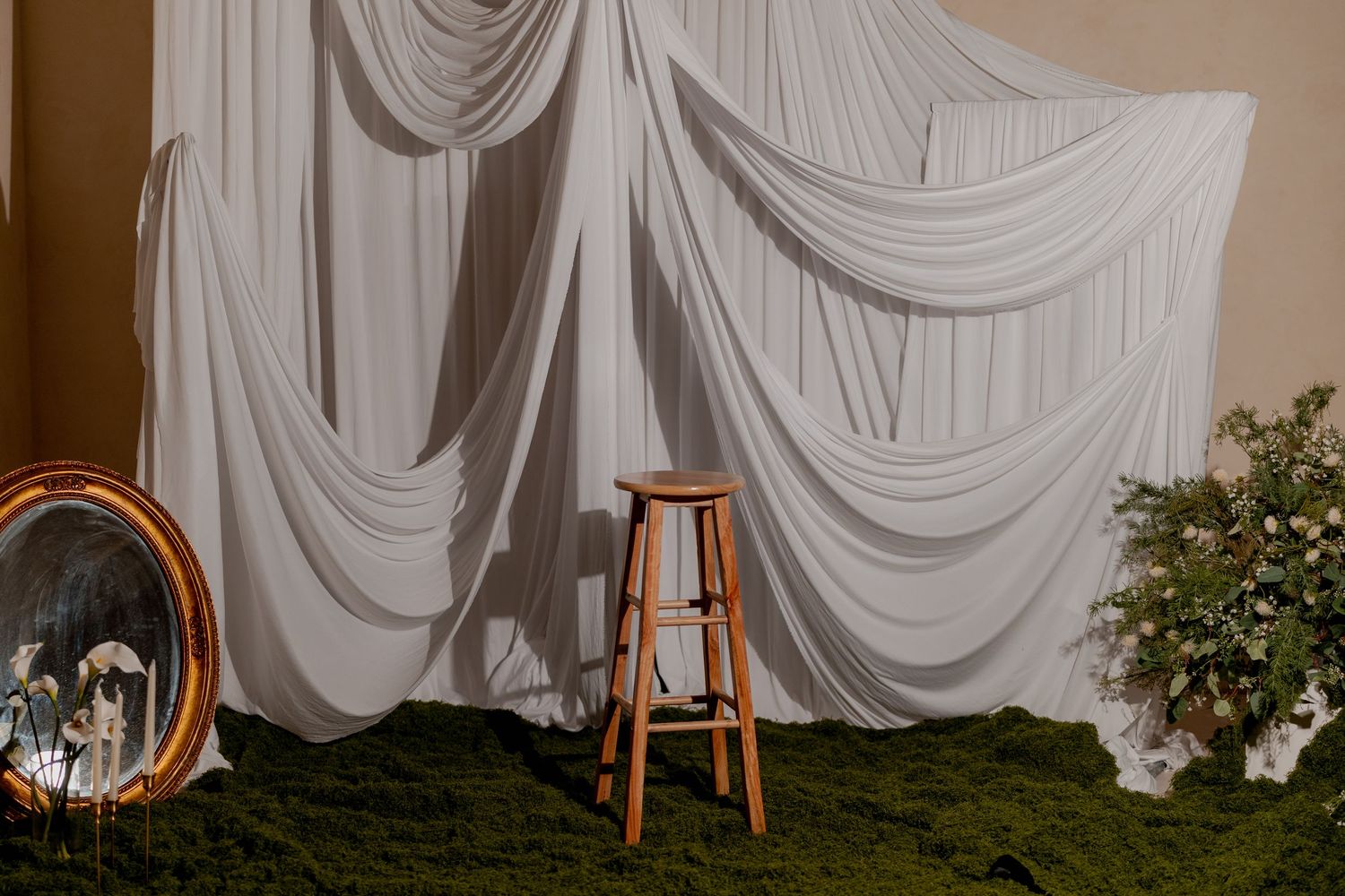 Elegant wedding backdrop with draped white fabric, wooden stool, moss ground cover, and floral arrangement on warm background.