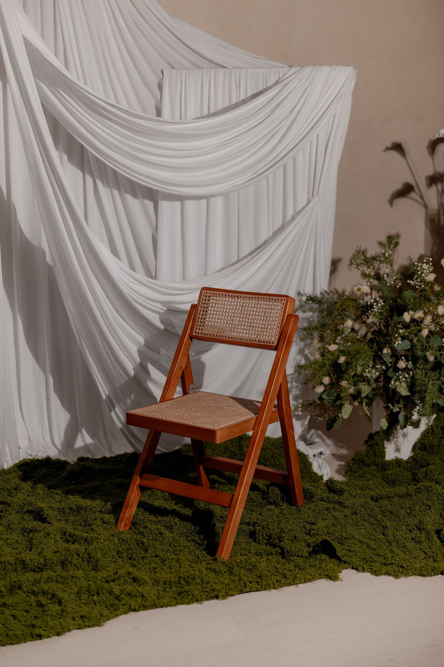 Wooden folding chair with rattan seat and back on green moss, white draped fabric backdrop, floral arrangement.