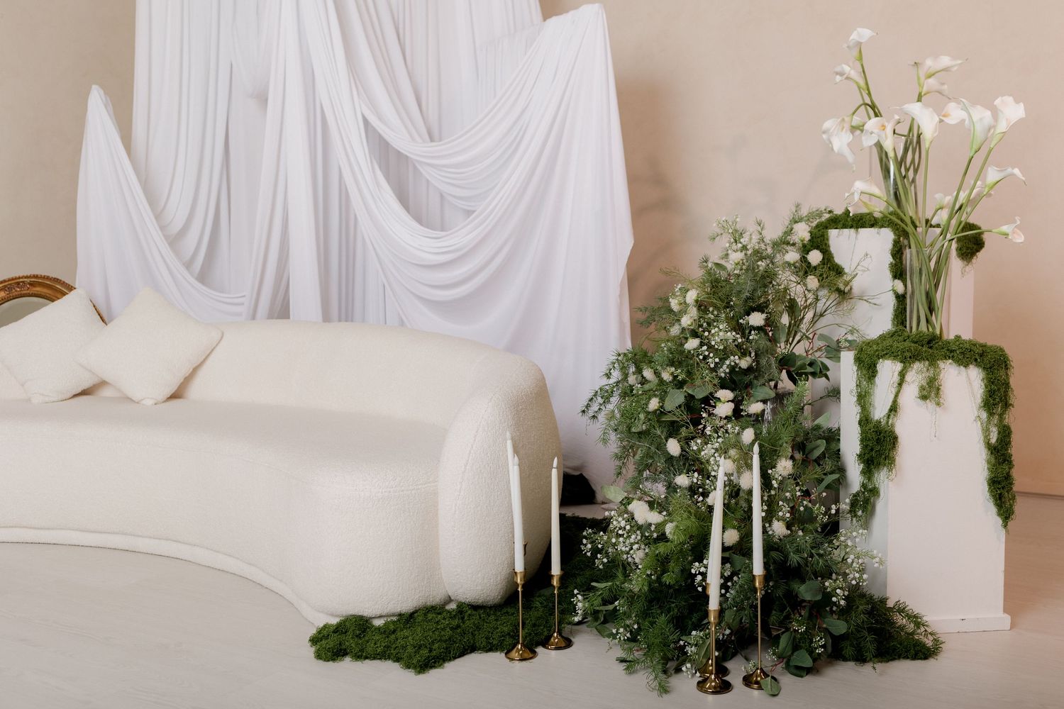 Elegant wedding setup with white draped backdrop, cream sofa, lush greenery, white floral arrangements, and gold candlesticks.