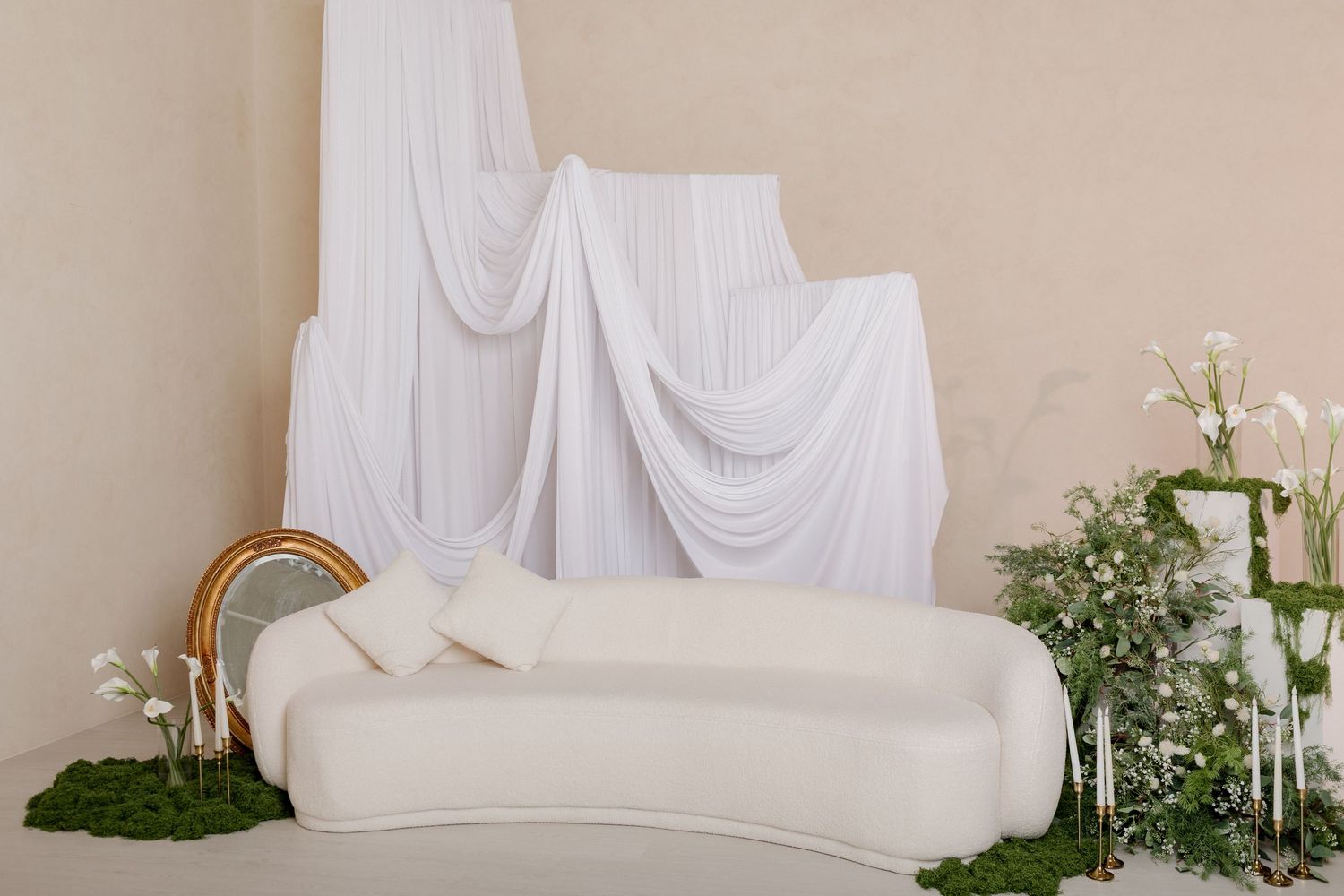 White curved sofa with draped fabric backdrop, gold mirror, and floral greenery arrangements in an elegant wedding setup.