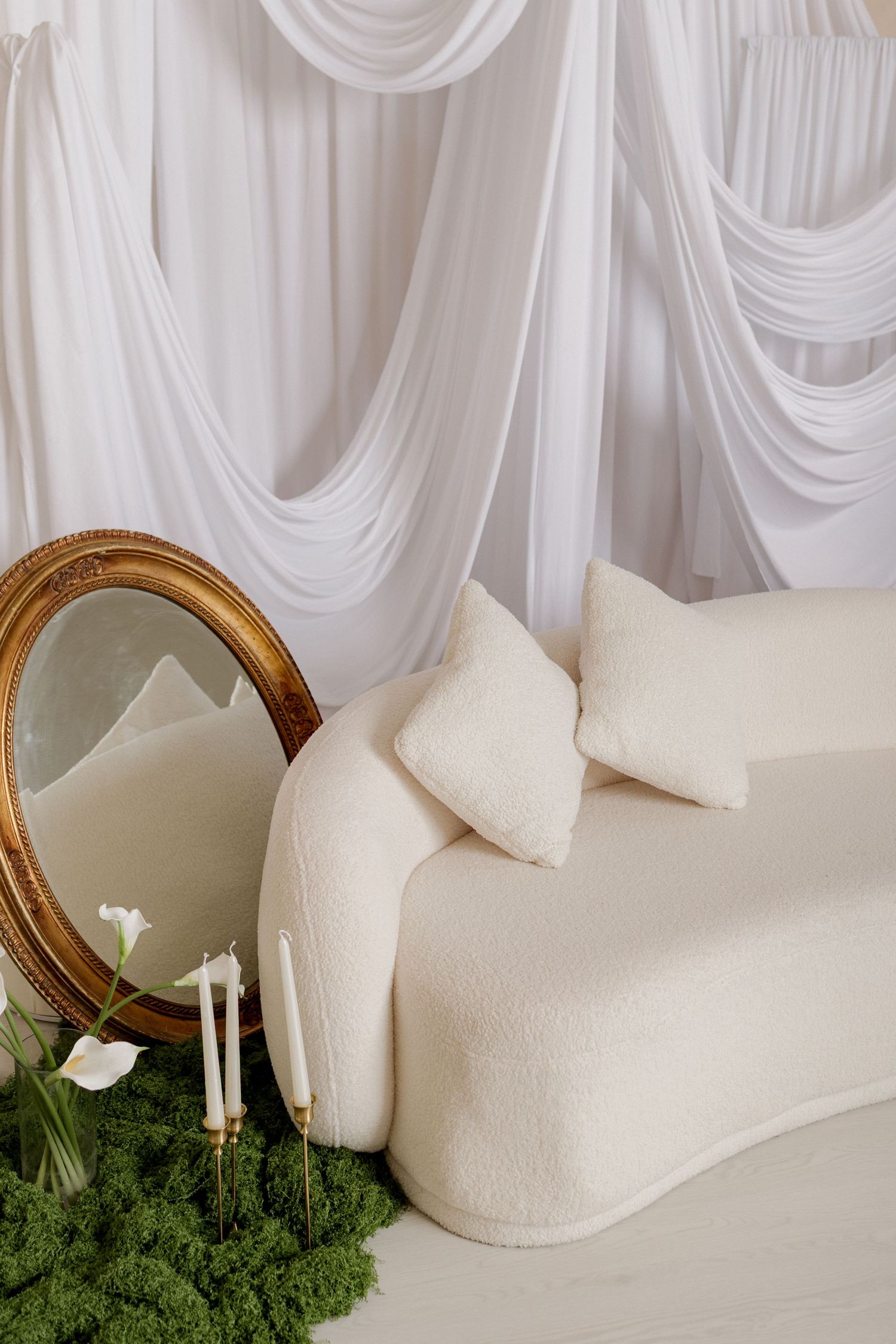 Elegant cream boucle curved sofa with pillows, gold oval mirror, white draping, candles, calla lilies, and green moss decor.