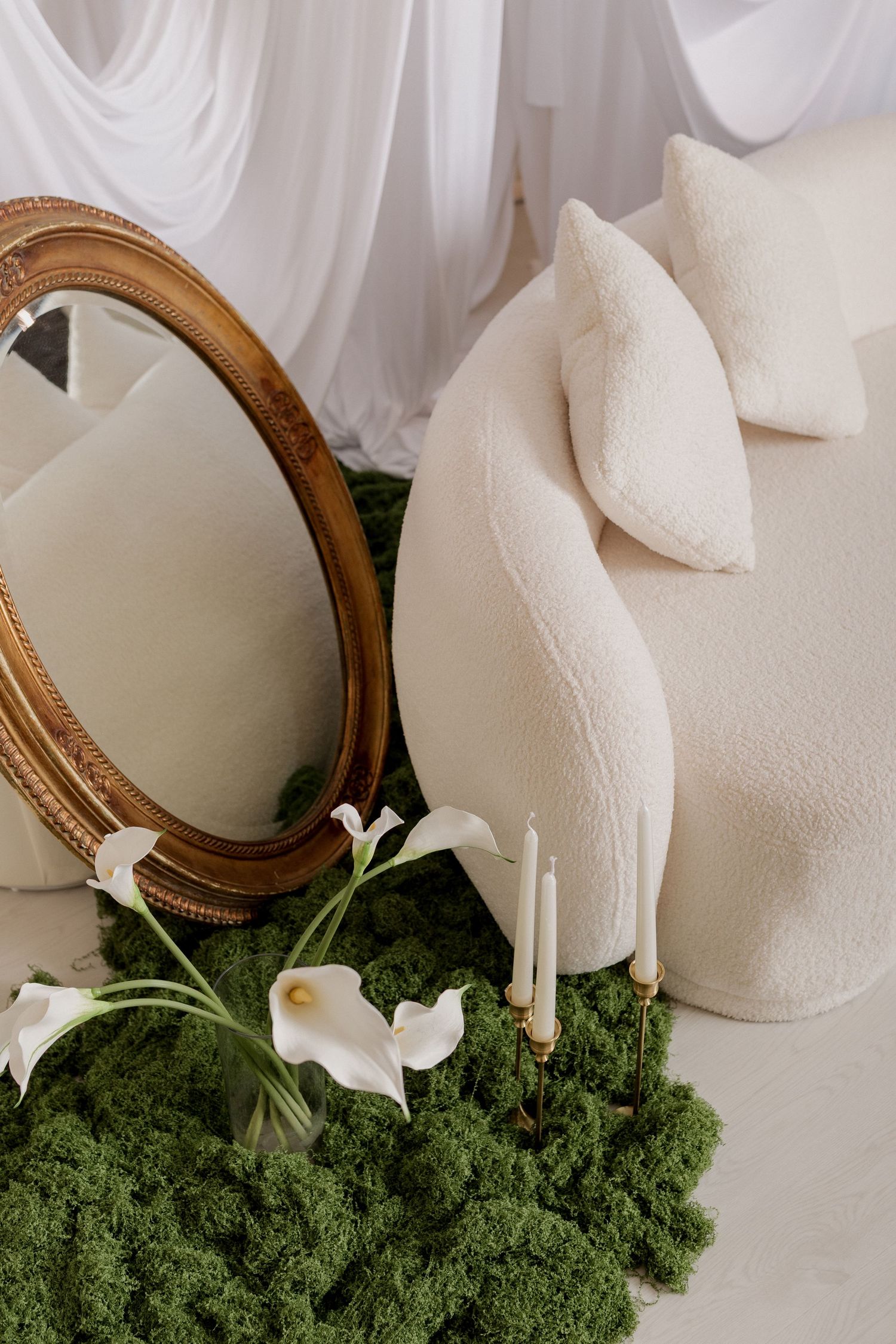 Elegant white bouclé sofa with pillows, gold oval mirror, calla lilies, candles, and green moss in a draped white room.