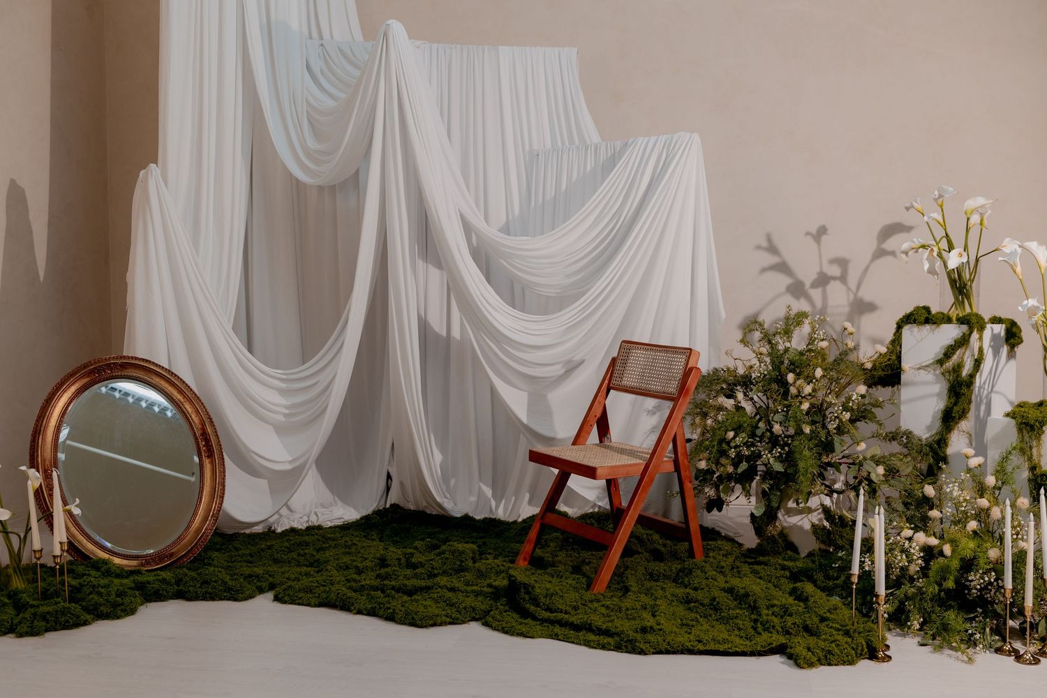 Elegant wedding backdrop with white draped fabric, wooden chair, oval mirror, green moss rug, and floral arrangements.
