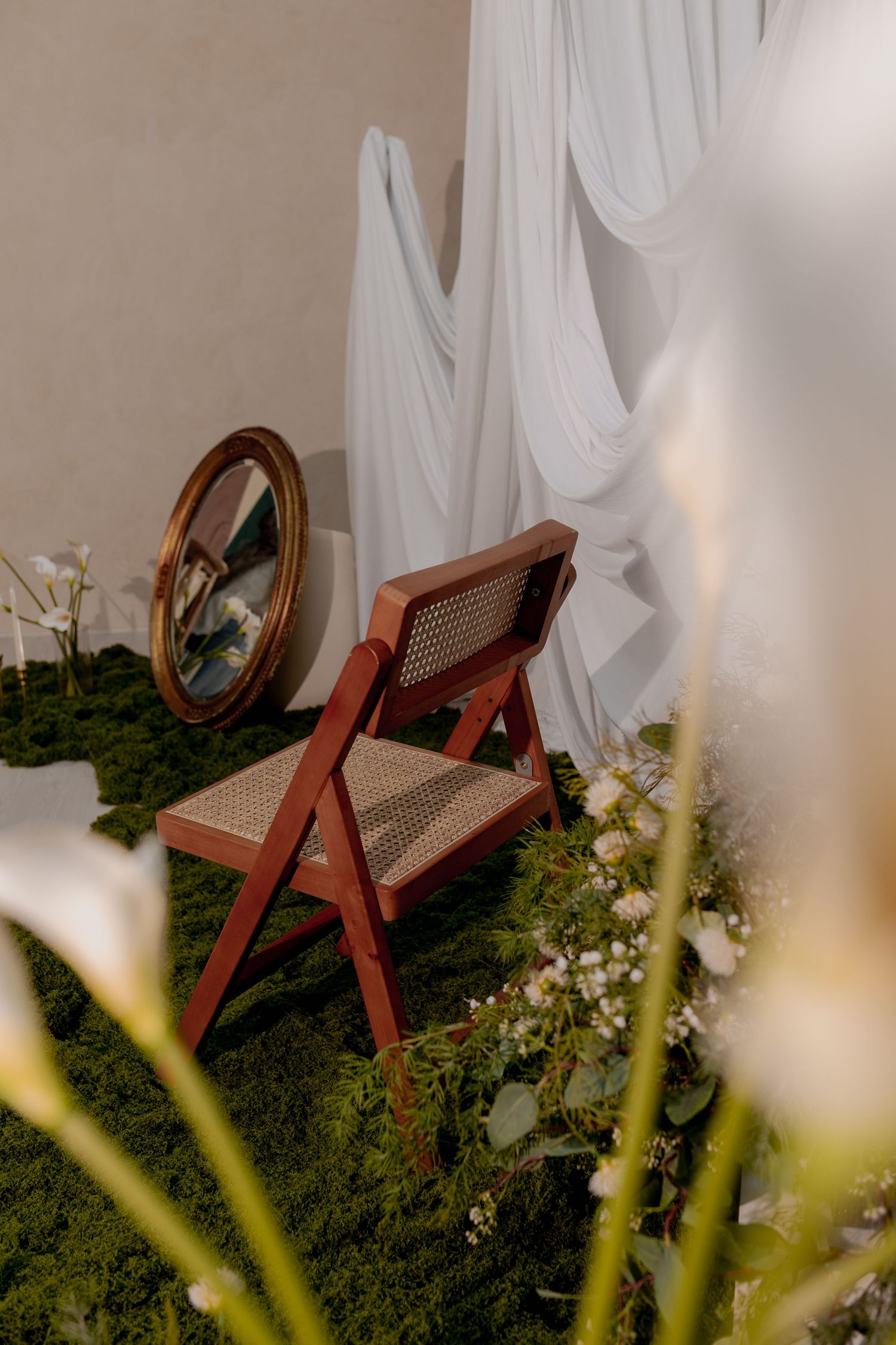 Rustic wooden folding chair with cane back on moss, beside oval mirror and white draped fabric backdrop.