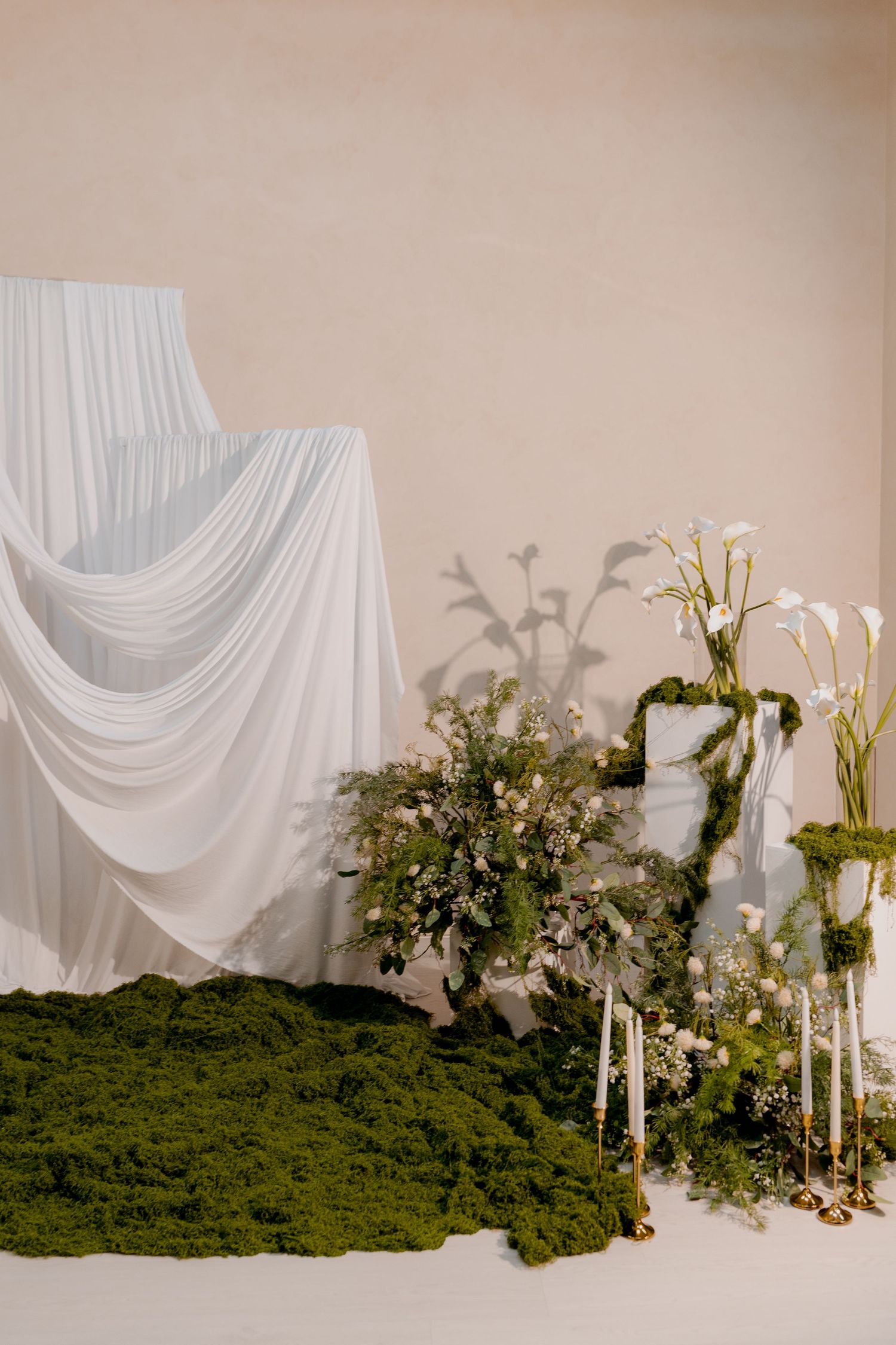 Elegant wedding backdrop with white draped fabric, green moss, floral arrangements, and tall candles on a neutral background.