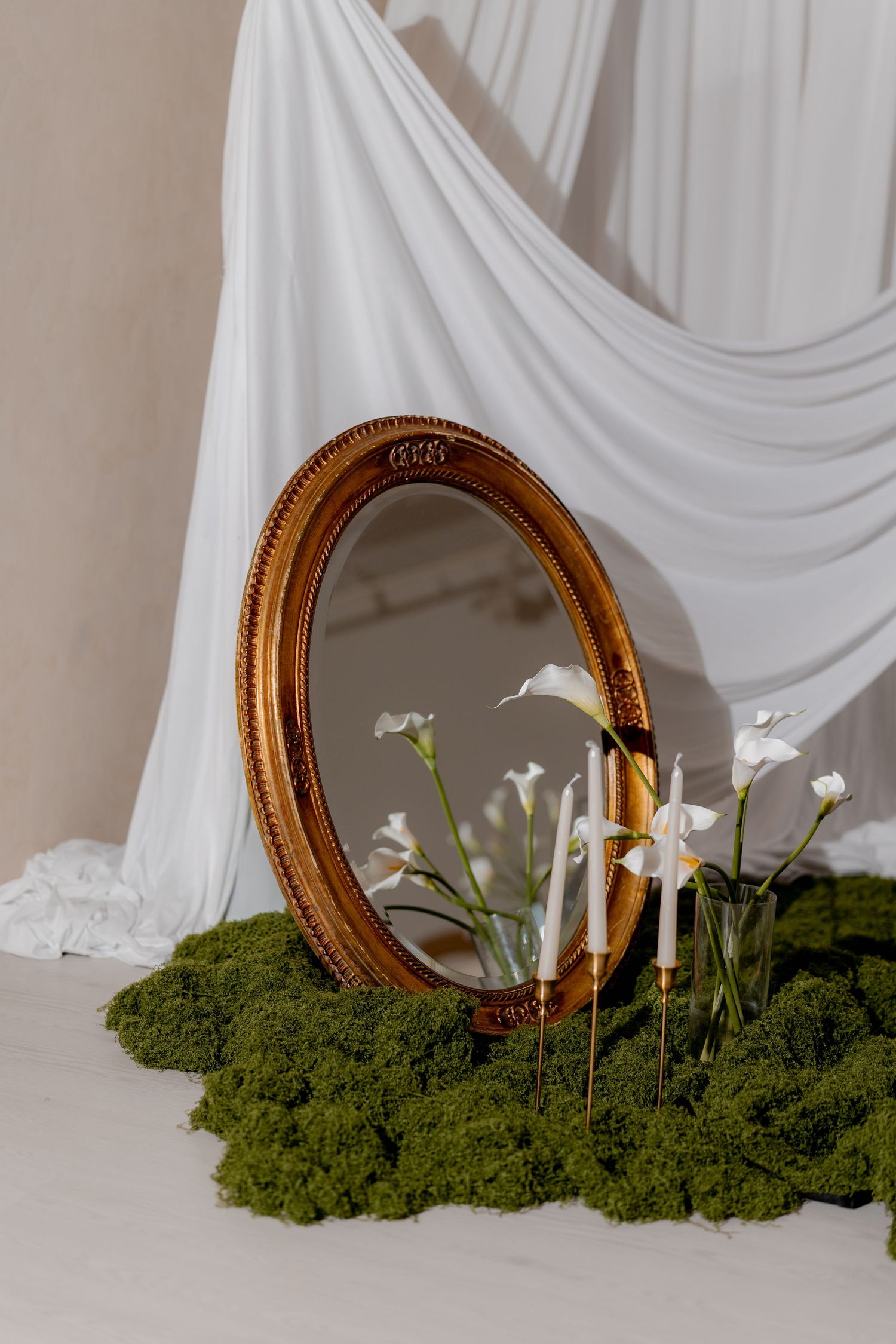 Gold oval mirror with white calla lilies and candles on green moss, draped white fabric backdrop.
