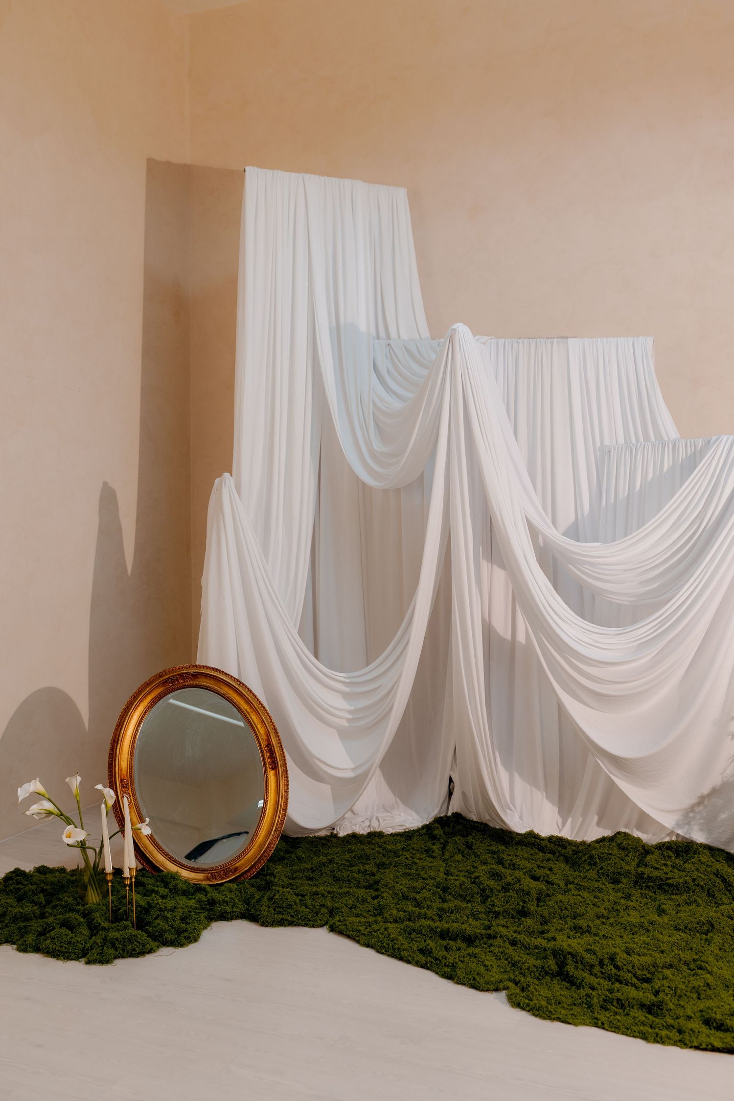Elegant white draped fabric backdrop with gold oval mirror and green moss floor decor in a beige studio setting.