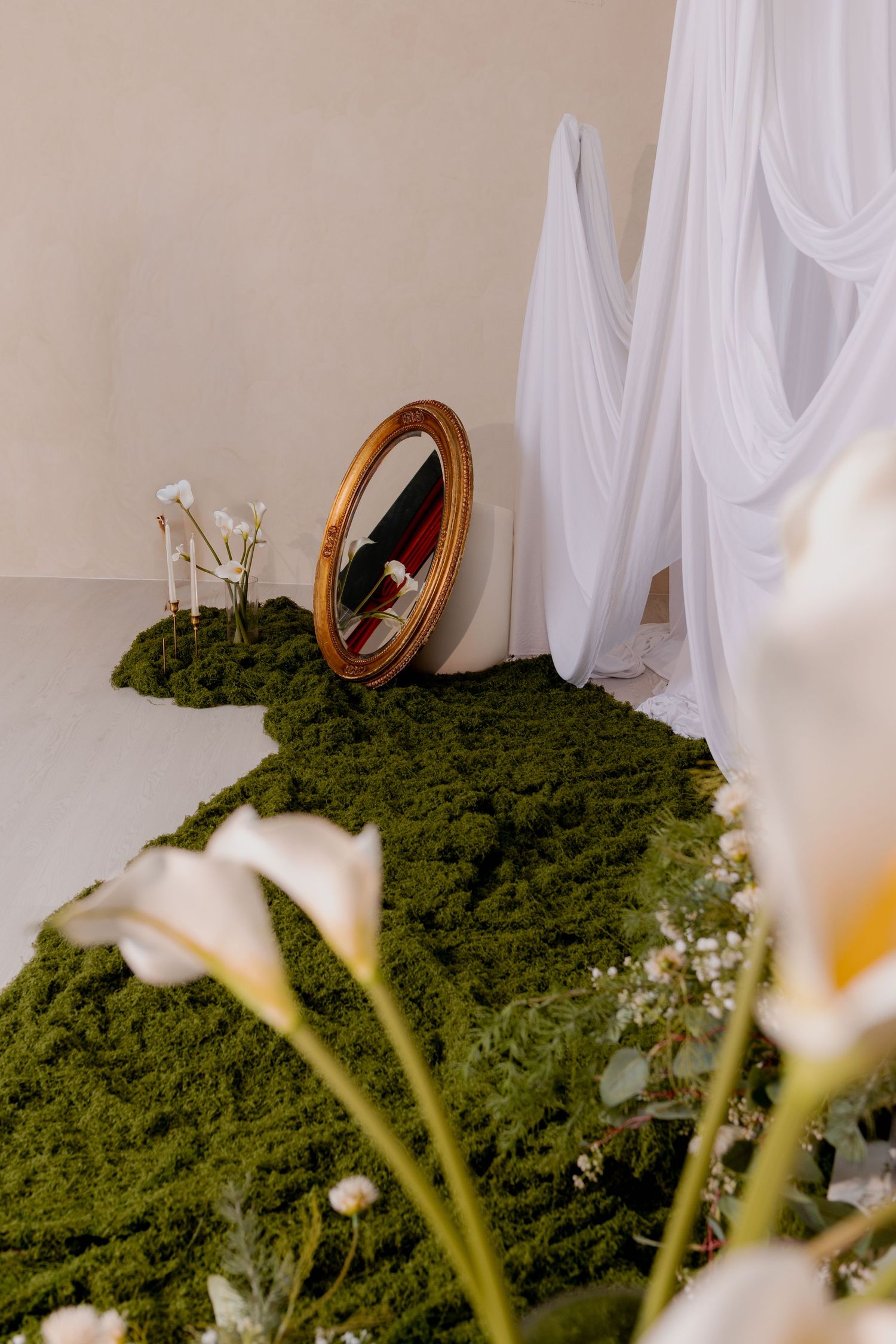 Oval gold mirror leaning against wall on green moss path with white calla lilies and flowing fabric.