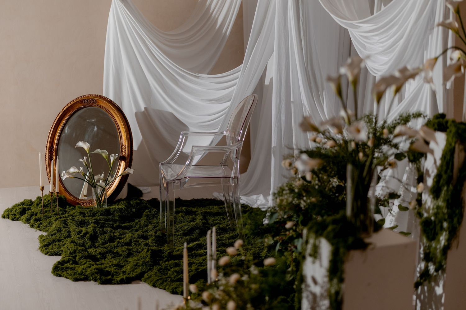 Elegant wedding altar with sheer white draping, ghost chair, gold oval mirror, moss greenery, and floral arrangements.