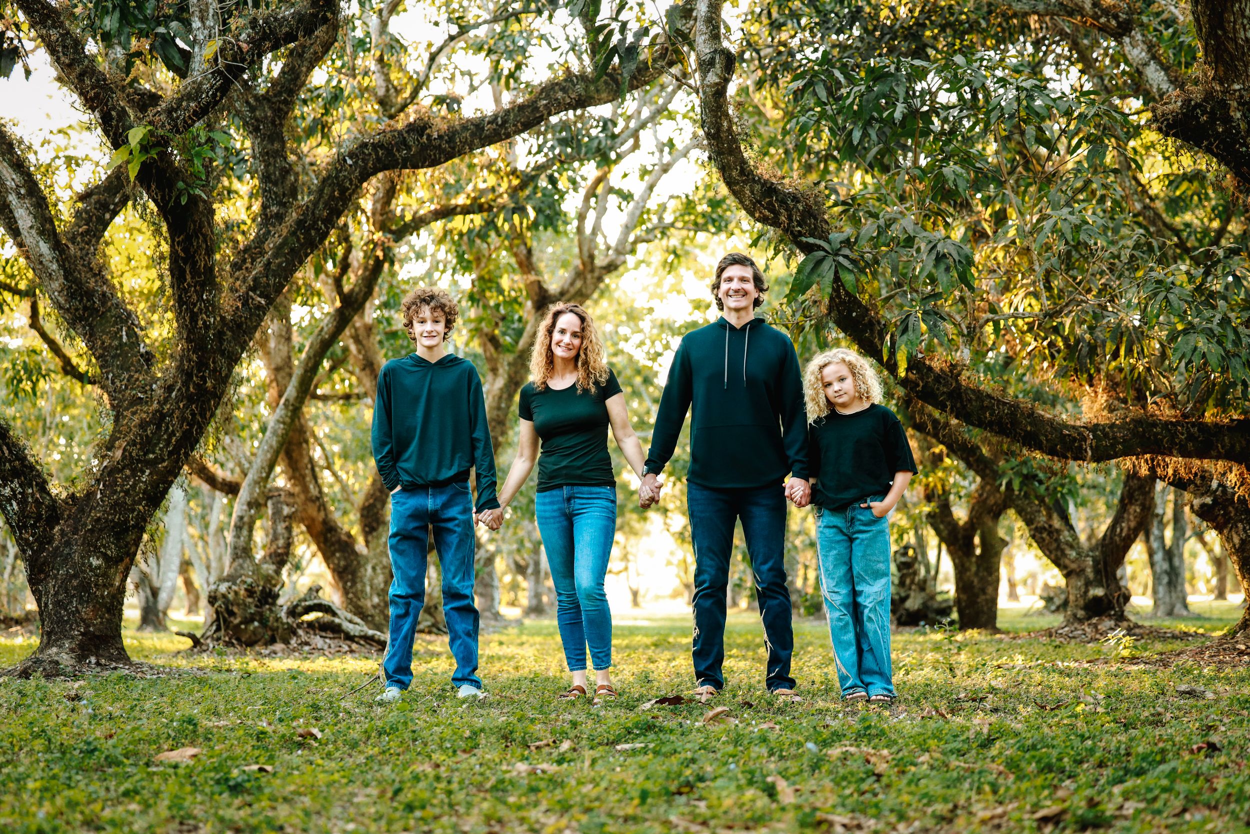 Family of four holding hands outdoors under large oak trees, wearing matching teal outfits in a lush green park.