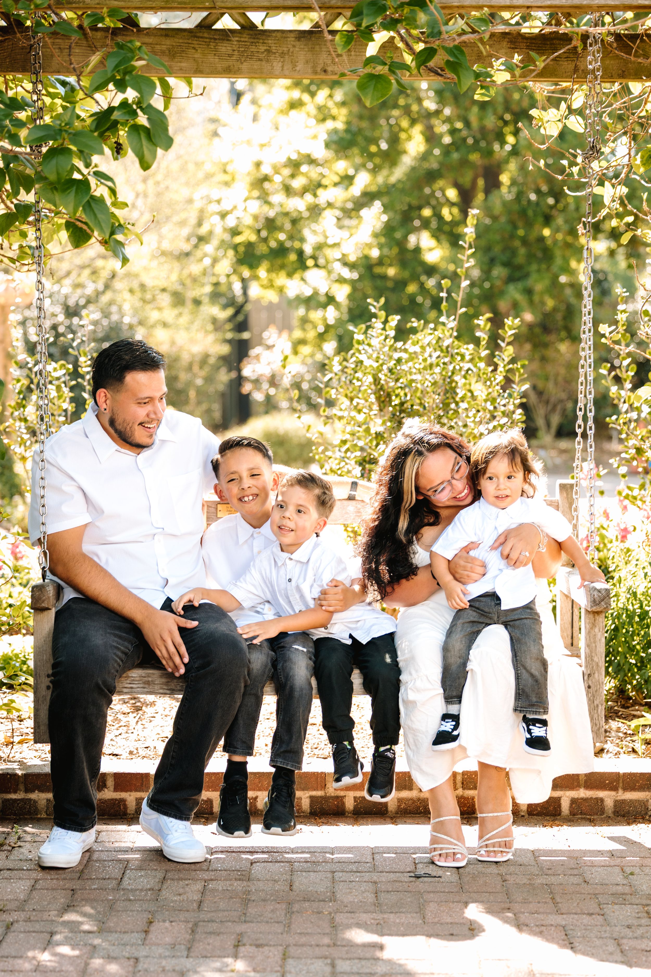 Happy family of five sitting on a garden swing bench, smiling outdoors under a pergola surrounded by lush greenery.
