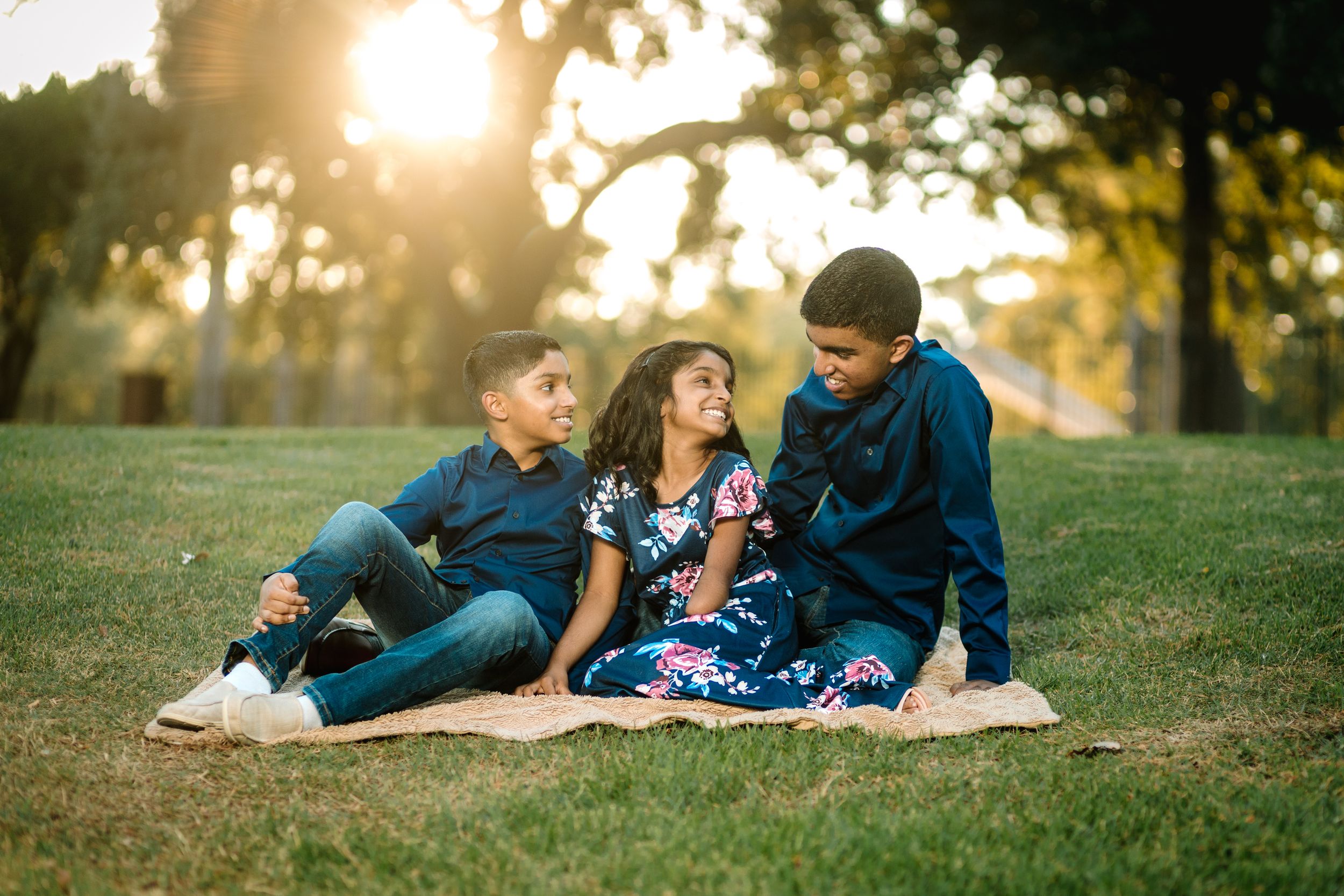 Three children sitting together on a blanket in a sunny park, smiling and enjoying quality time outdoors.