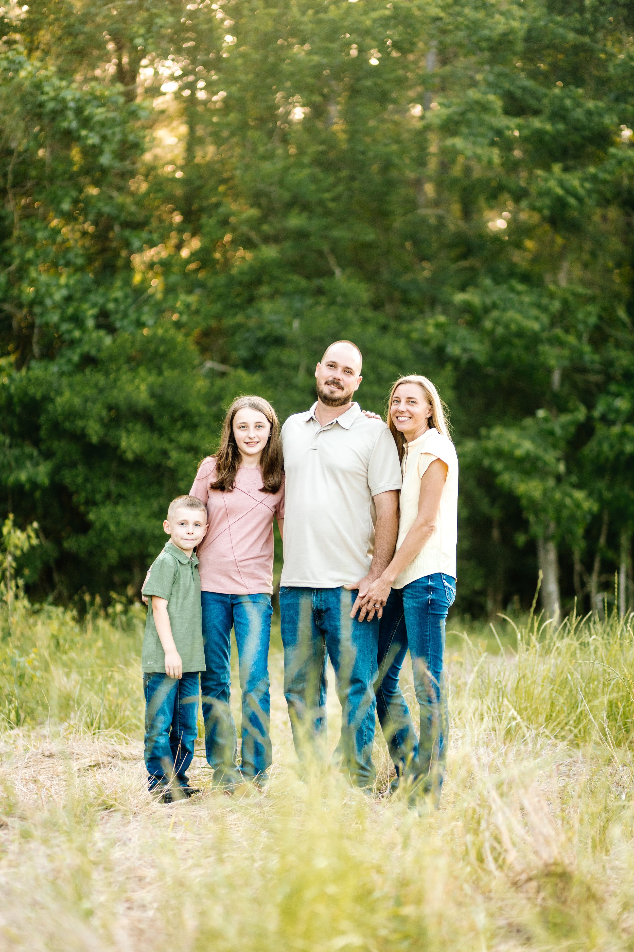 Family of four posing outdoors in a green meadow, smiling together in casual jeans and pastel shirts.