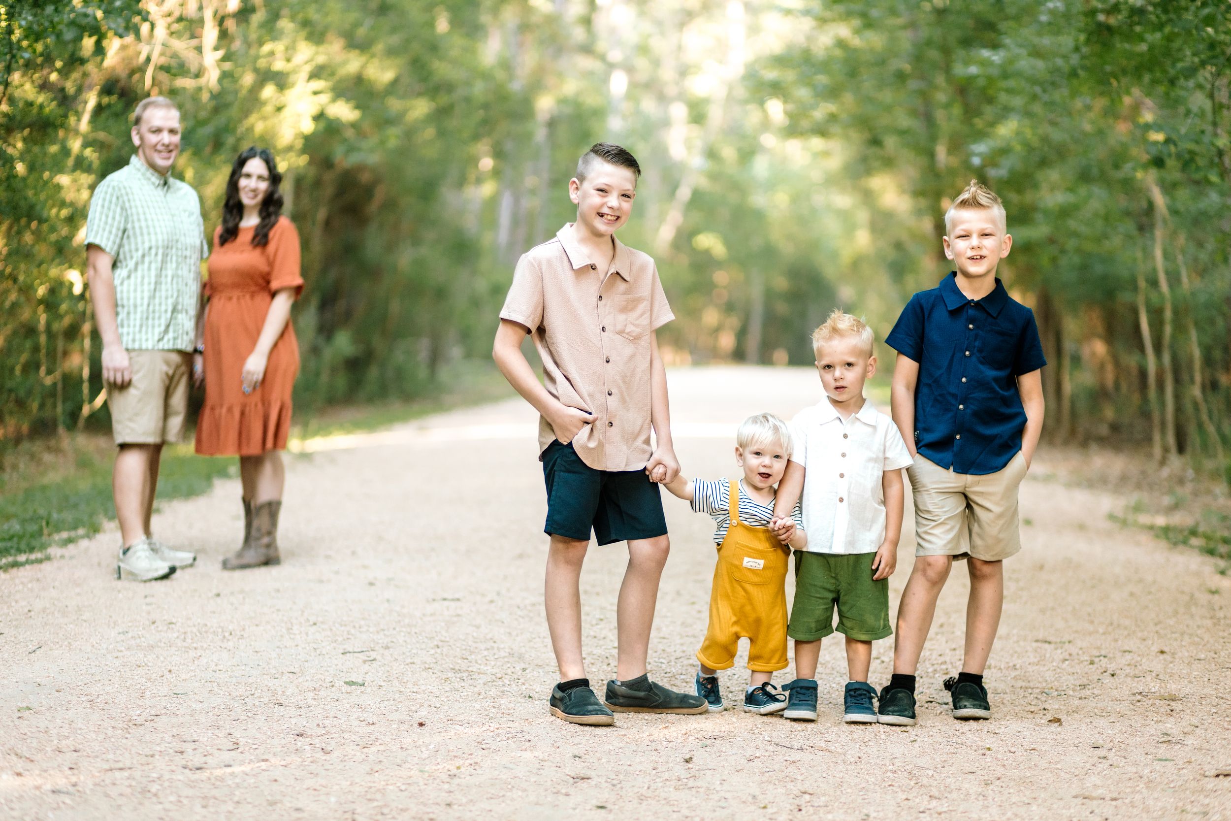 Family of five posing on a scenic tree-lined dirt path, parents standing behind three young boys smiling outdoors.