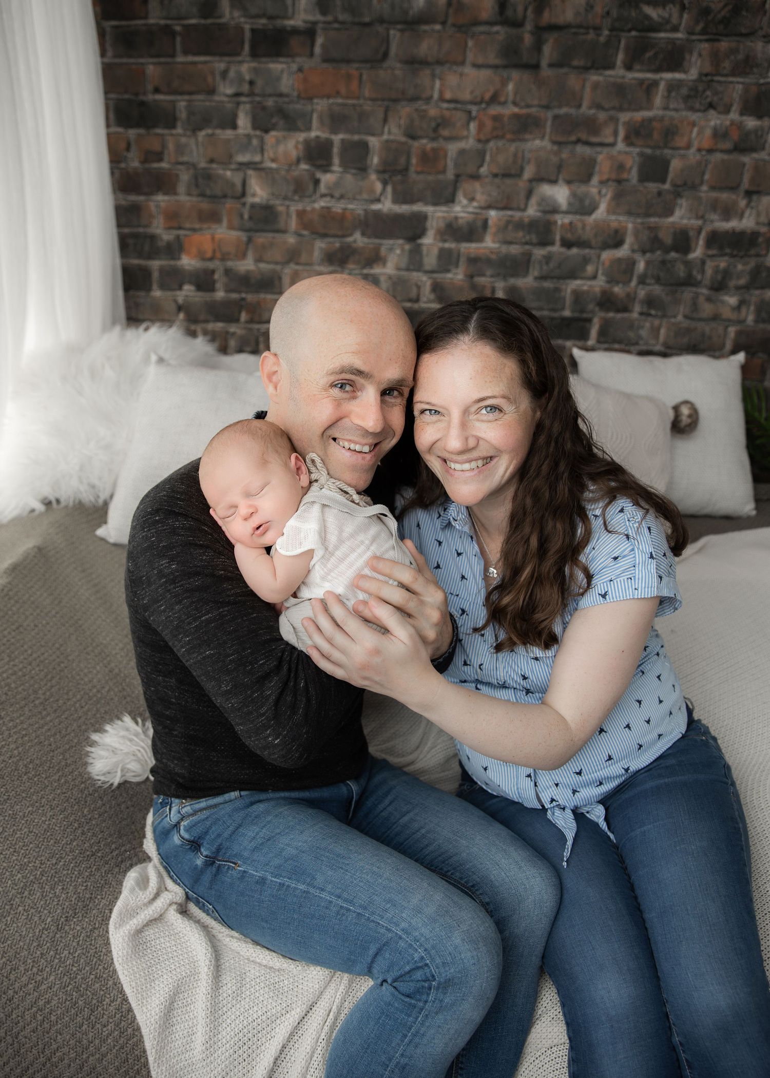 Happy couple sitting on a bed holding their newborn baby, smiling warmly in front of a rustic brick wall. Newborn Photography Cork