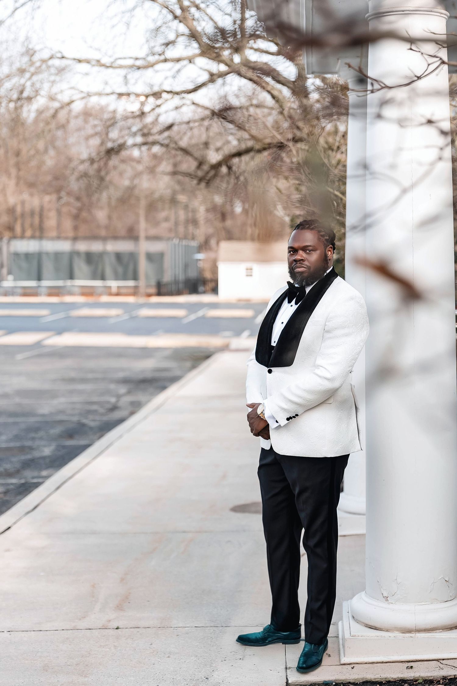 A well-dressed Black man in a white tuxedo jacket and black bow tie leans against a white column outdoors. Deerfield Golf Club Newark De