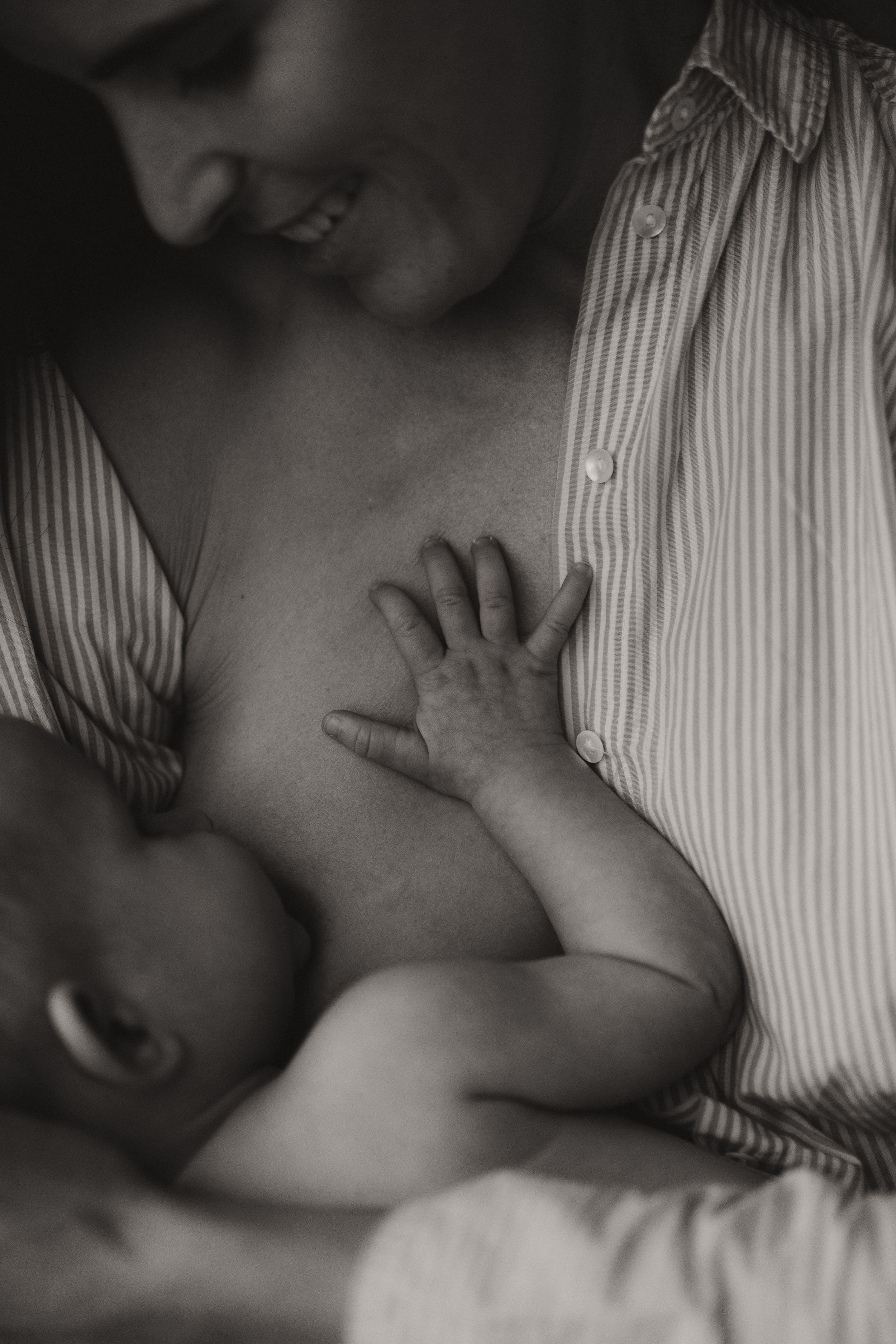 Black and white photo of a smiling mother breastfeeding her newborn baby, with tiny hand resting on her chest.