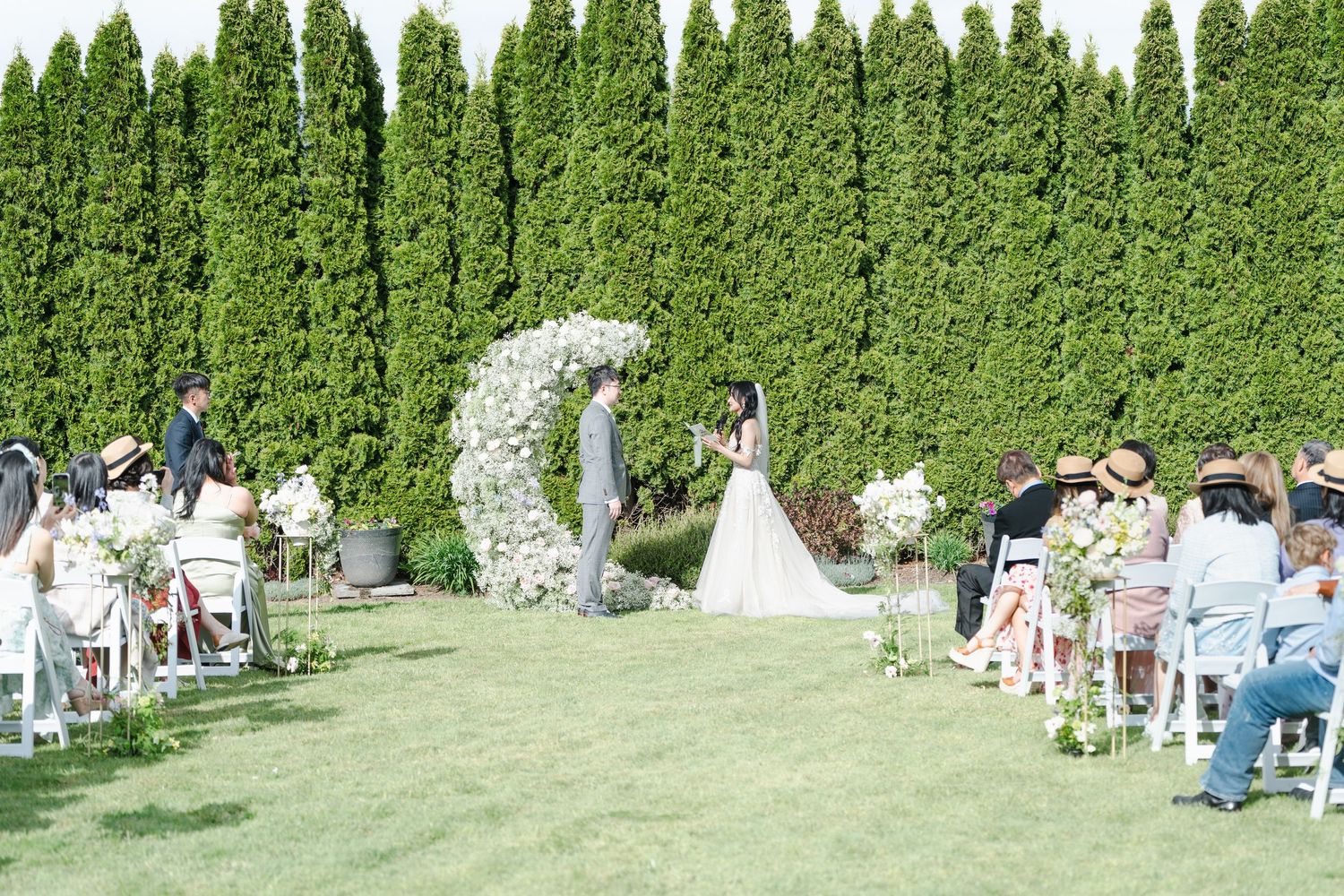 Outdoor wedding ceremony on green lawn with floral arch, bride and groom, guests seated along aisle, tall hedges backdrop.
