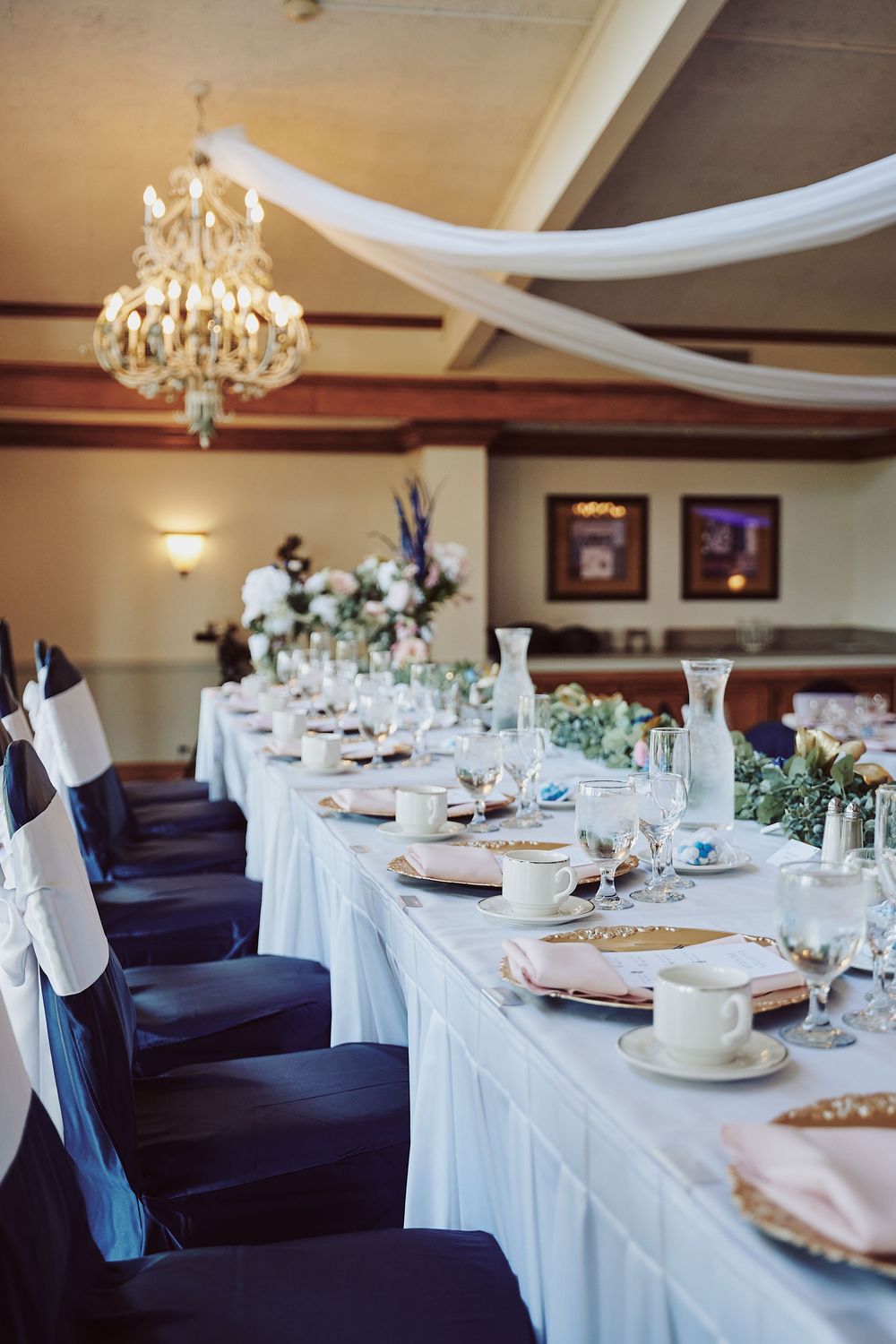 Elegant wedding reception hall with long banquet table, navy chair covers, crystal chandelier, white draping, and floral centerpieces.