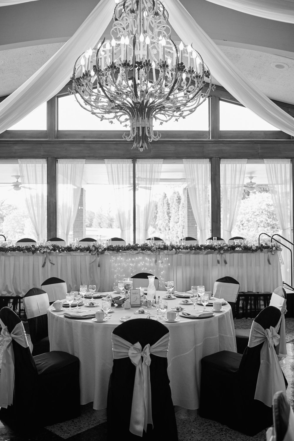 Elegant black and white wedding reception with chandelier, draped ceilings, floral garland, and chairs with white sashes.
