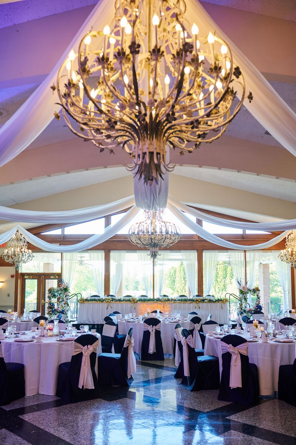 Elegant wedding reception hall with ornate chandelier, draped white fabric ceiling, and tables set with black chair covers and pink sashes.