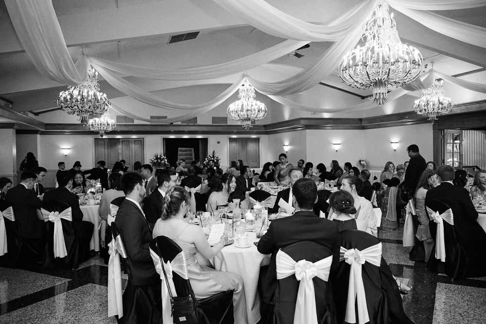Black and white photo of an elegant wedding reception in a ballroom with chandeliers, draped ceilings, and guests seated at round tables.