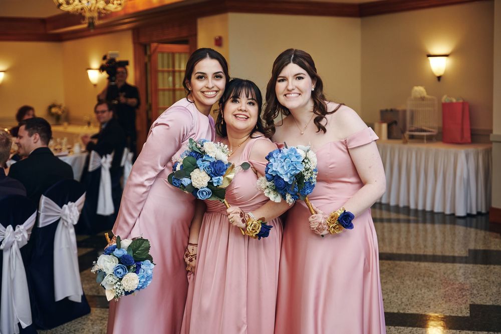 Three bridesmaids in pink off-shoulder gowns holding blue and white bouquets pose together at an elegant wedding reception.