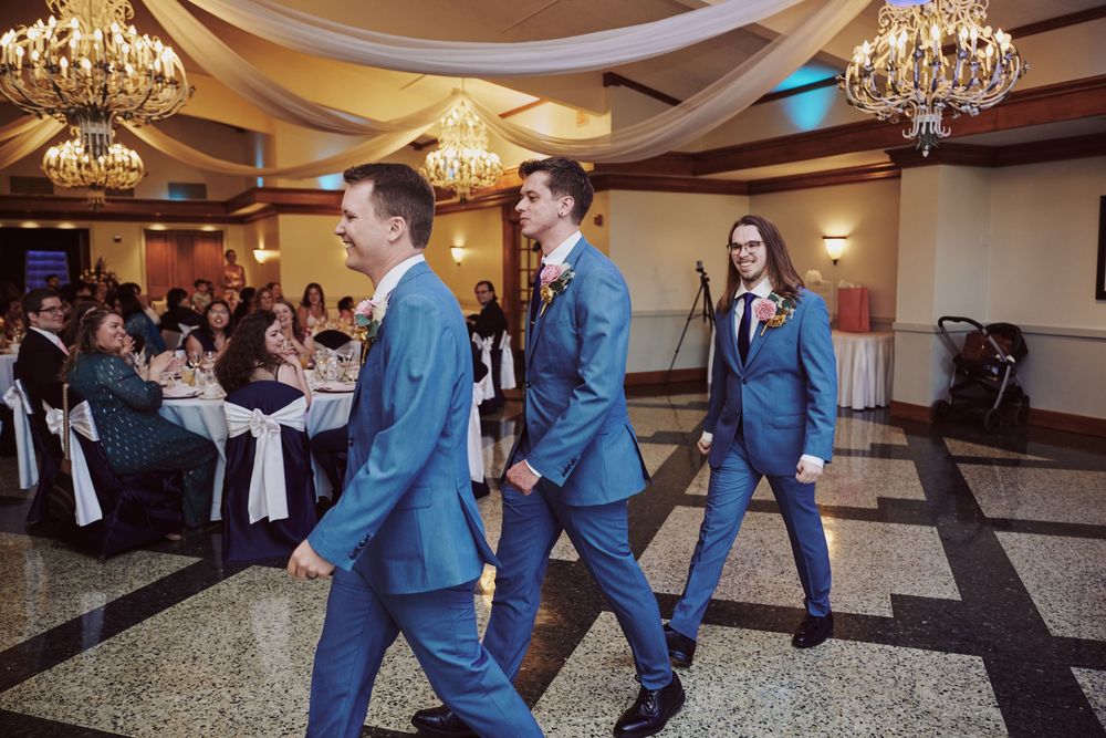 Three groomsmen in blue suits walking across a wedding reception ballroom with chandeliers and guests seated at tables.