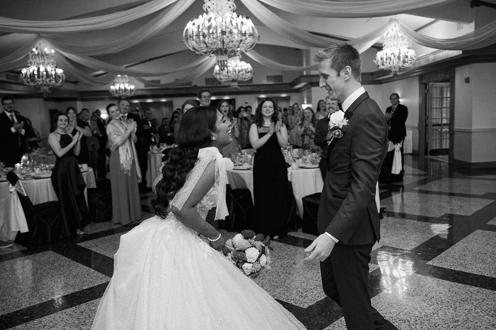 Bride and groom share their first dance at a grand wedding reception with chandeliers and guests applauding in black and white.
