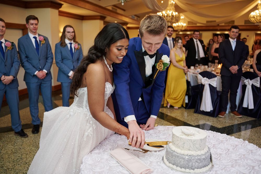 Bride and groom cutting wedding cake at elegant reception hall with groomsmen in blue suits watching.