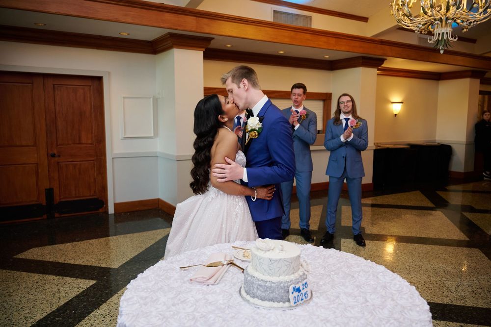 Bride and groom share a kiss while cutting their wedding cake as groomsmen in blue suits applaud nearby.