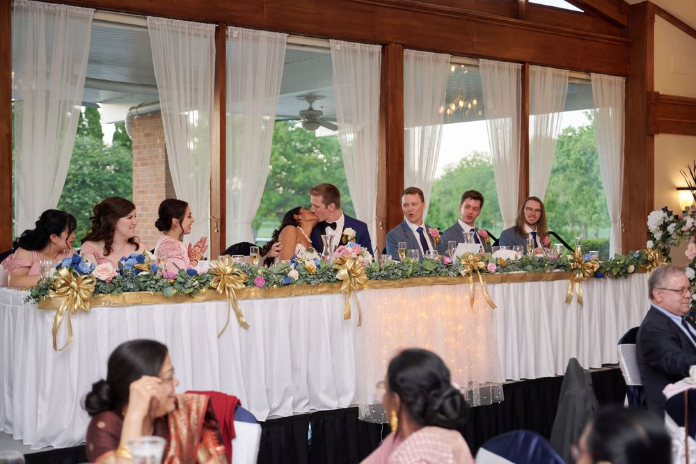 Wedding party seated at head table during reception, adorned with floral arrangements, as couple shares a kiss.