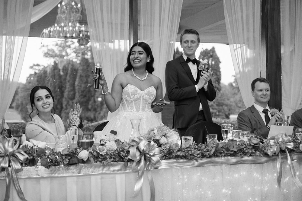 Black and white wedding photo of bride and groom toasting with guests at elegantly decorated head table.