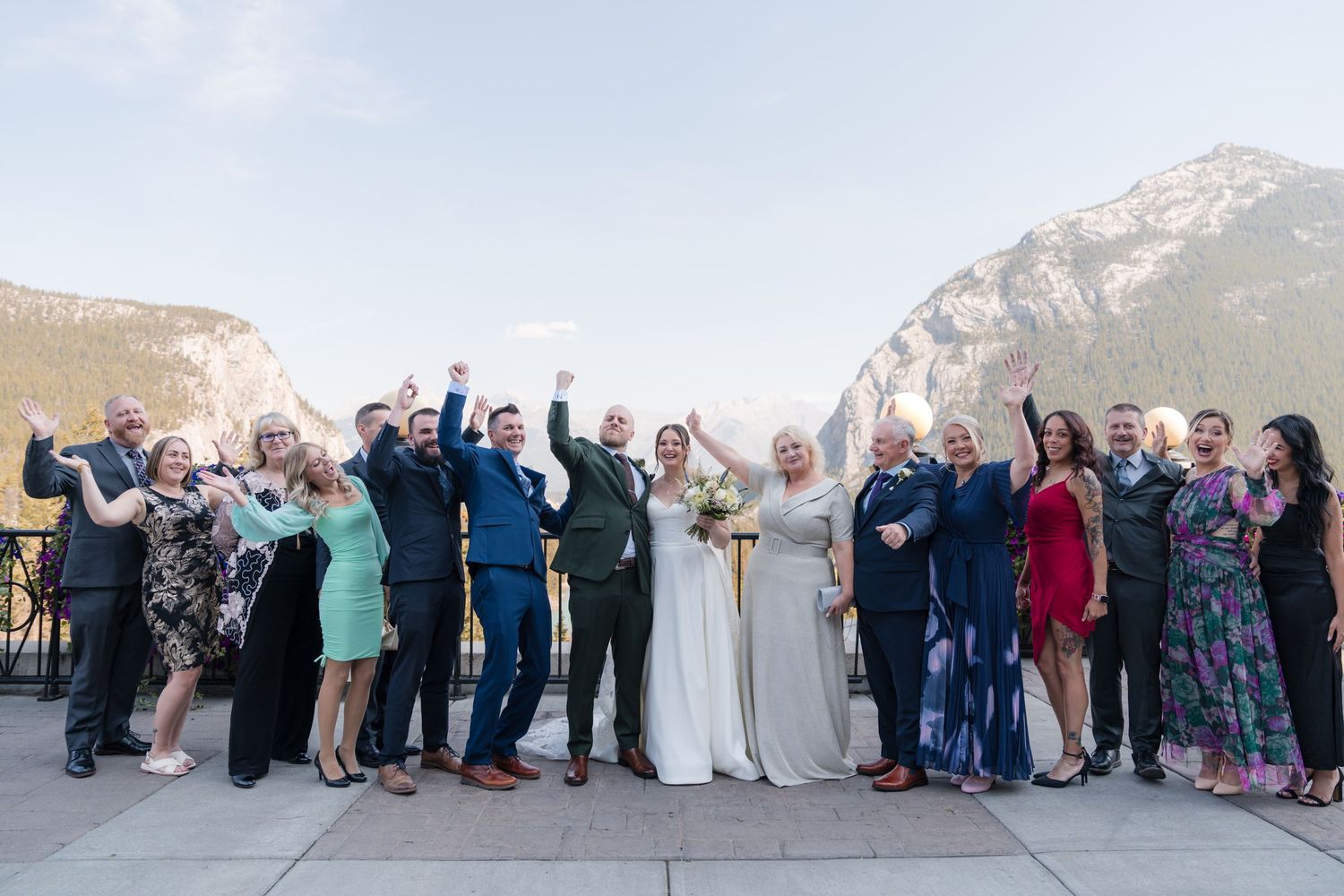 Wedding party celebrating outdoors with mountains in background, guests cheering with arms raised at the Farimont Banff Springs Hotel.