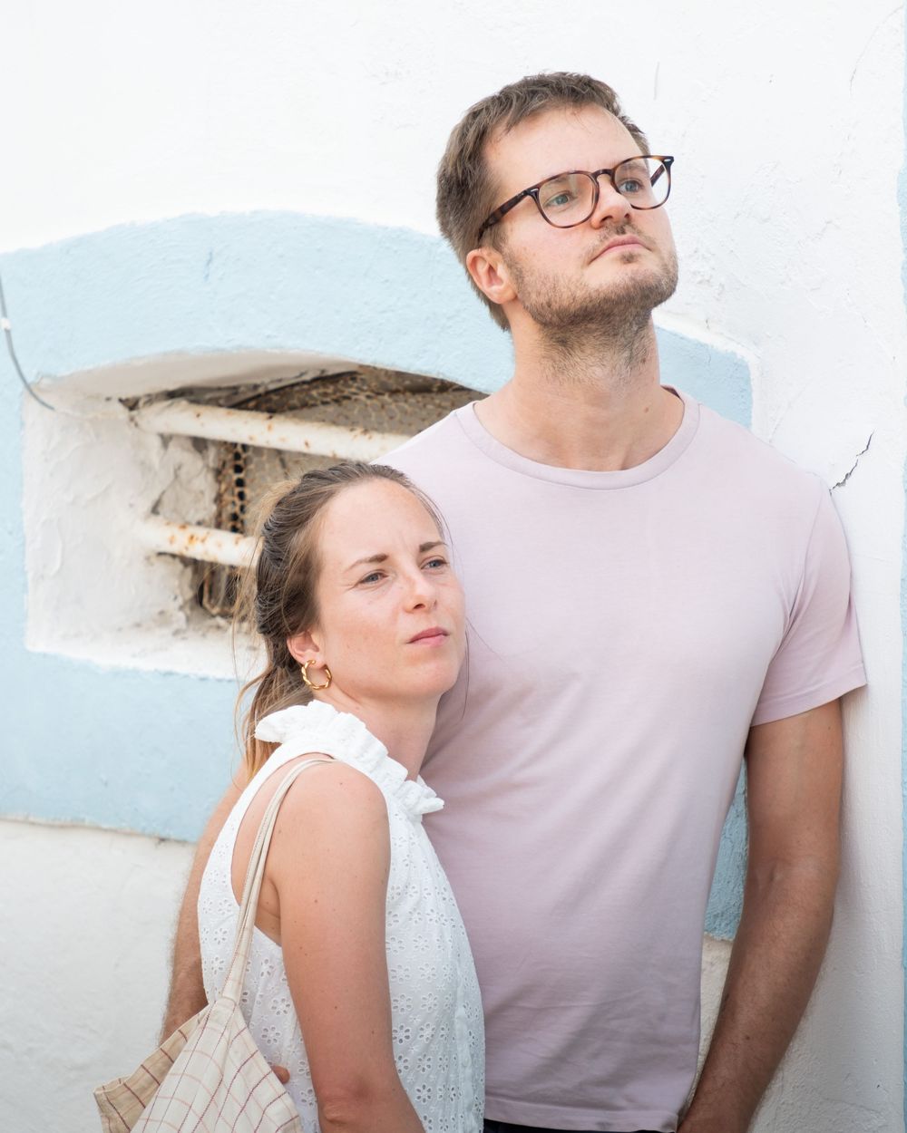 Couple posing against a white Mediterranean wall, woman in white dress, man in pink t-shirt with glasses.