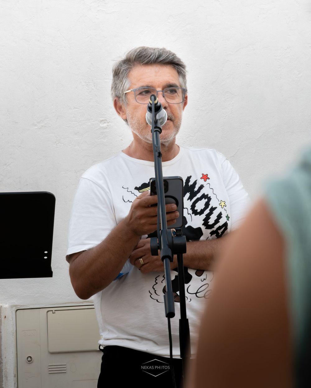 Middle-aged man with gray hair holding a microphone stand, wearing a white graphic t-shirt indoors.
